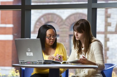 Two female students sitting at a table with an opened laptop in a university room, discussing their school work.