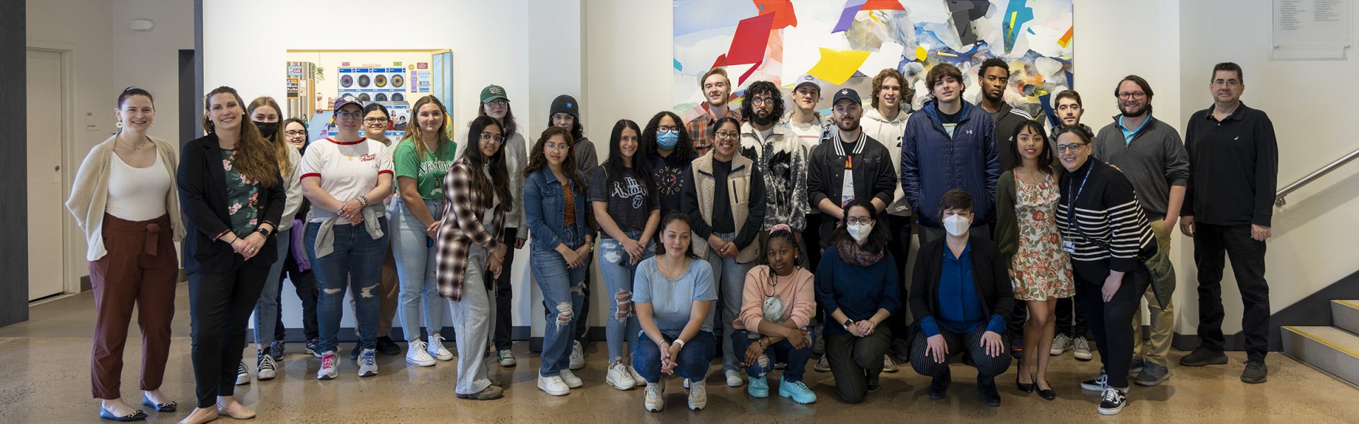 A group photo of UConn Waterbury students from the Urban Studies Class during their field trip to the MAT. The group of students and their instructors are standing in front of the wall with a colorful painting in the main entrance of the museum.