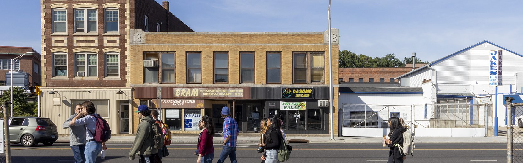 UConn Waterbury students from the Urban Studies Class walking across the street in front of Dabomb Salad shop, Jake's Autoshop, and BRAM commercial restaurant equipment shop in downtown Waterbury.